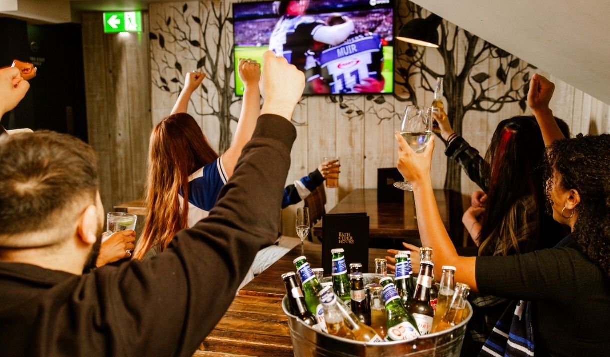 people cheering around a table watching rugby, with a bucket of beer bottles on the table